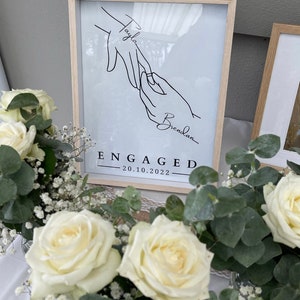May include: A framed print with a minimalist line drawing of two hands, one with a ring, and the names "Tayla" and "Brendan." Below, the word "ENGAGED" and the date "20.10.2022" are displayed. The frame is light wood.