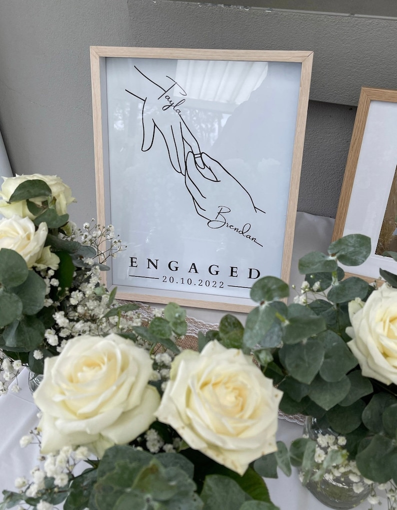 May include: A framed print with a minimalist line drawing of two hands, one with a ring, and the names "Tayla" and "Brendan." Below, the word "ENGAGED" and the date "20.10.2022" are displayed. The frame is light wood.