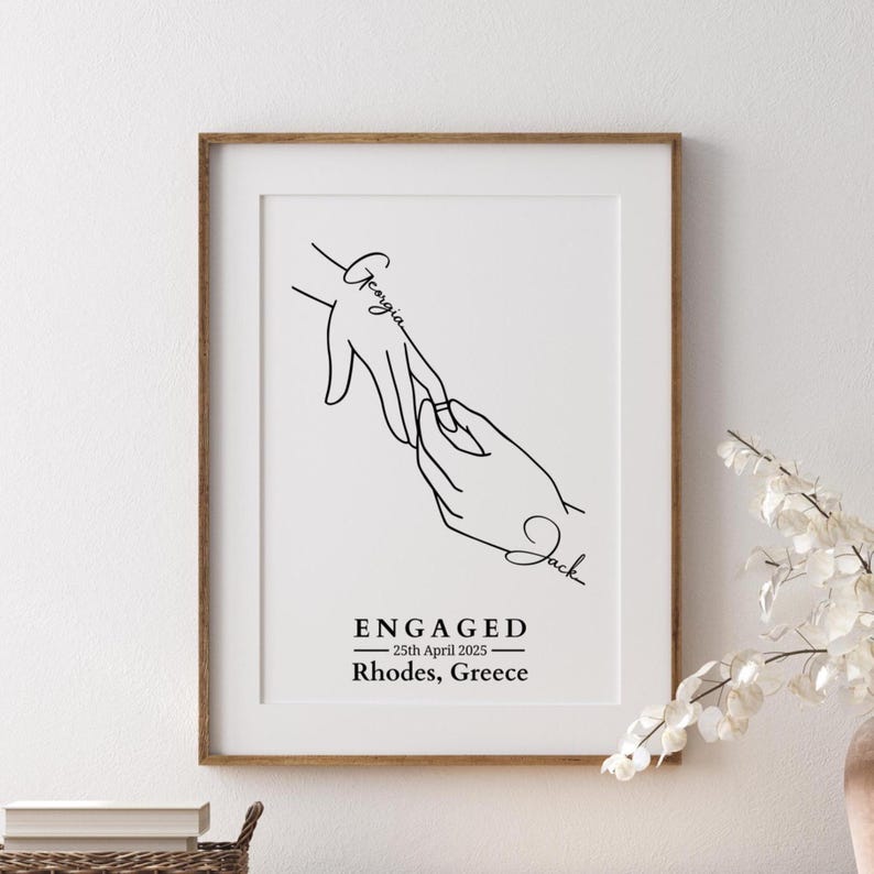 May include: A framed black and white line art print. The artwork depicts two hands, one placing a ring on the other's finger. The names "Georgia" and "Jack" are written on the hands. The word "ENGAGED" and the date "25th April 2025" are below the hands, along with "Rhodes, Greece."