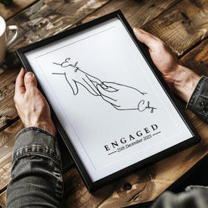 May include: A framed black and white print featuring a line drawing of two hands, one placing a ring on the other's finger. The names "Emily" and "Coby" are above the hands, with "ENGAGED - 25th December 2025" below.