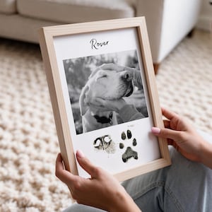 Op de afbeelding: Een ingelijste zwart-witfoto van een hond die geaaid wordt, met de naam "Rover" erboven. Onder de foto staan twee afdrukken van poten. De lijst is van lichtgekleurd hout.