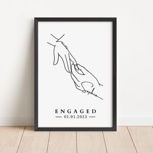 May include: Black framed art print with a minimalist line drawing of two hands, one placing a ring on the other. The names "Lucas" and "Sophia" are written on the hands. The word "ENGAGED" and the date "01.01.2023" are below the hands.