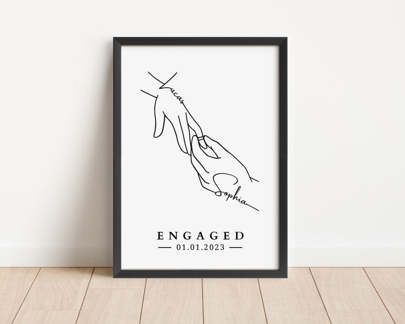 May include: Black framed art print with a minimalist line drawing of two hands, one placing a ring on the other. The names "Lucas" and "Sophia" are written on the hands. The word "ENGAGED" and the date "01.01.2023" are below the hands.