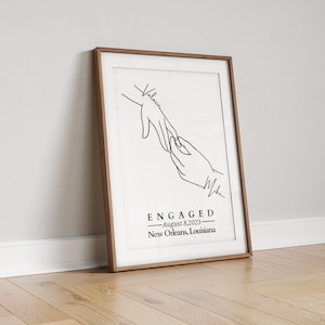 May include: A framed art print with a minimalist line drawing of two hands, one reaching for the other. The word "ENGAGED" is centered below the hands, with the date "August 8, 2023" and location "New Orleans, Louisiana" below it. The names "Valarie" and "Mike" are written on the print.