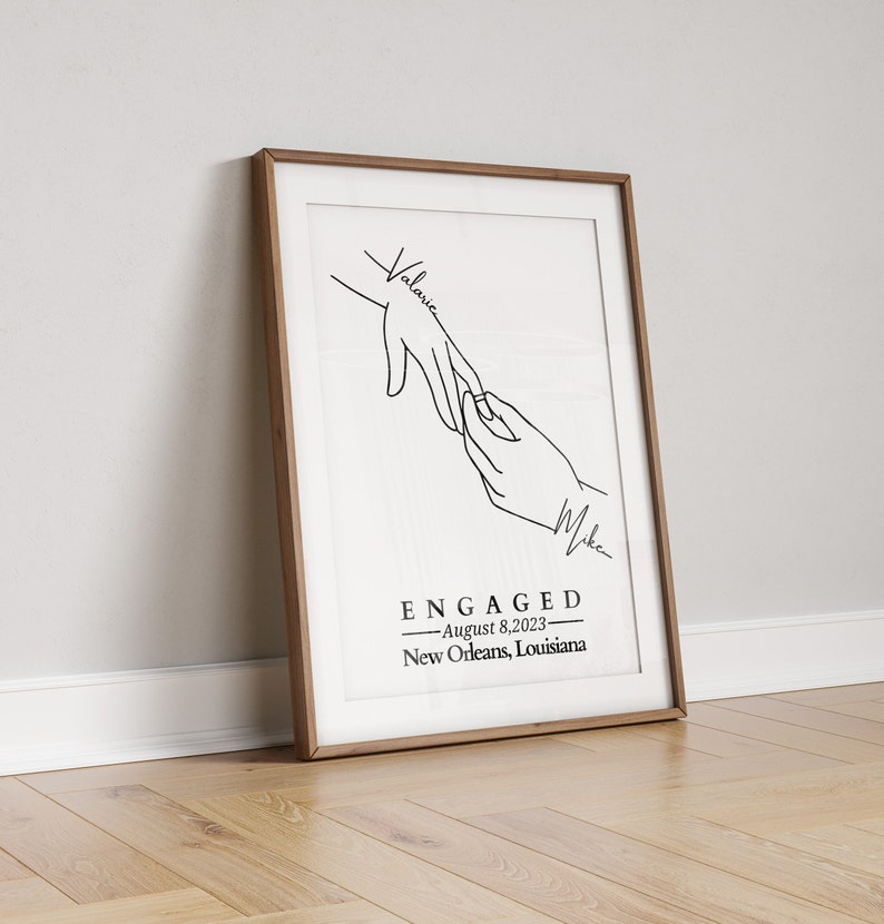May include: A framed art print with a minimalist line drawing of two hands, one reaching for the other. The word "ENGAGED" is centered below the hands, with the date "August 8, 2023" and location "New Orleans, Louisiana" below it. The names "Valarie" and "Mike" are written on the print.