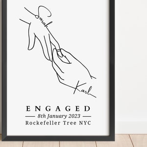 May include: Black and white line art print featuring two hands, one placing a ring on the other. The names "Sarah" and "Karl" are written on the hands. The word "ENGAGED" and the date "8th January 2023" are also included.