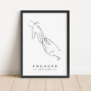 May include: A framed black and white print featuring a line drawing of two hands, one placing a ring on the other. The word "Kirsten" is written on one hand, and "Cocray" on the other. The word "ENGAGED" and the date "28.01.2023" are printed below.