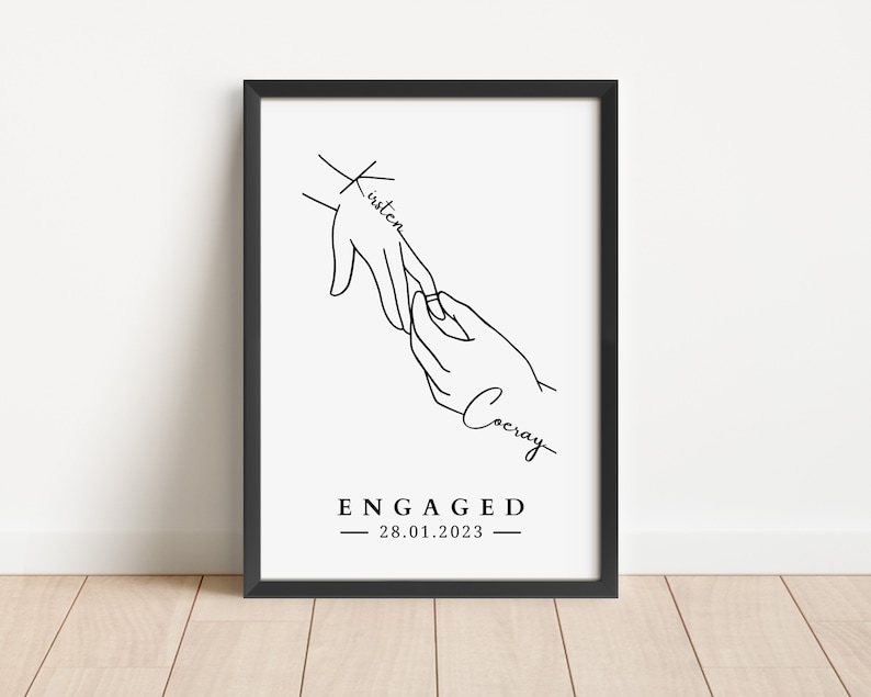 May include: A framed black and white print featuring a line drawing of two hands, one placing a ring on the other. The word "Kirsten" is written on one hand, and "Cocray" on the other. The word "ENGAGED" and the date "28.01.2023" are printed below.