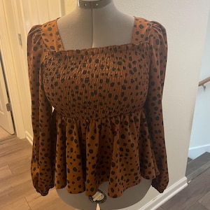 May include: A brown long-sleeved top with a square neckline and a peplum hem. The top has a smocked bodice and is patterned with black spots. The garment is displayed on a mannequin.