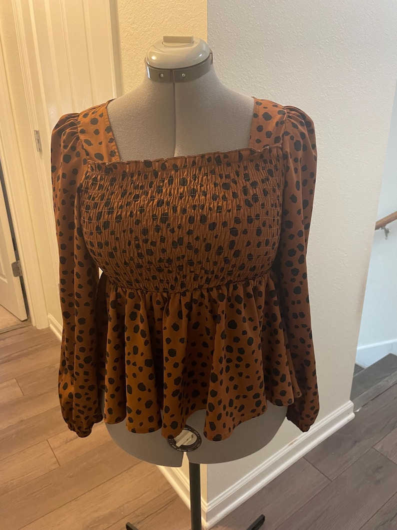 May include: A brown long-sleeved top with a square neckline and a peplum hem. The top has a smocked bodice and is patterned with black spots. The garment is displayed on a mannequin.