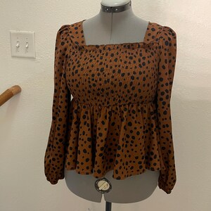 May include: A brown long-sleeved top with a square neckline and a peplum hem. The top has a smocked bodice and is covered in black spots. The sleeves are long and gathered at the cuffs. The top is displayed on a mannequin.