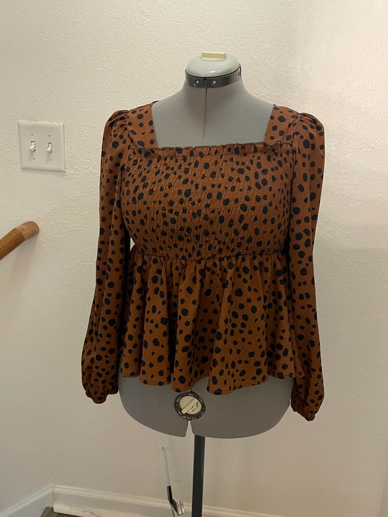May include: A brown long-sleeved top with a square neckline and a peplum hem. The top has a smocked bodice and is covered in black spots. The sleeves are long and gathered at the cuffs. The top is displayed on a mannequin.