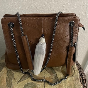 May include: A brown leather handbag with a chevron pattern and a silver chain strap. The bag has a rectangular shape with a zip closure and a small handle. A tag is attached to the side of the bag.