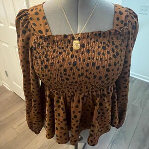 May include: A brown top with black polka dots, a square neckline, and long sleeves. The top has a smocked bodice and a peplum hem. A gold-coloured necklace with a square pendant hangs on the mannequin.