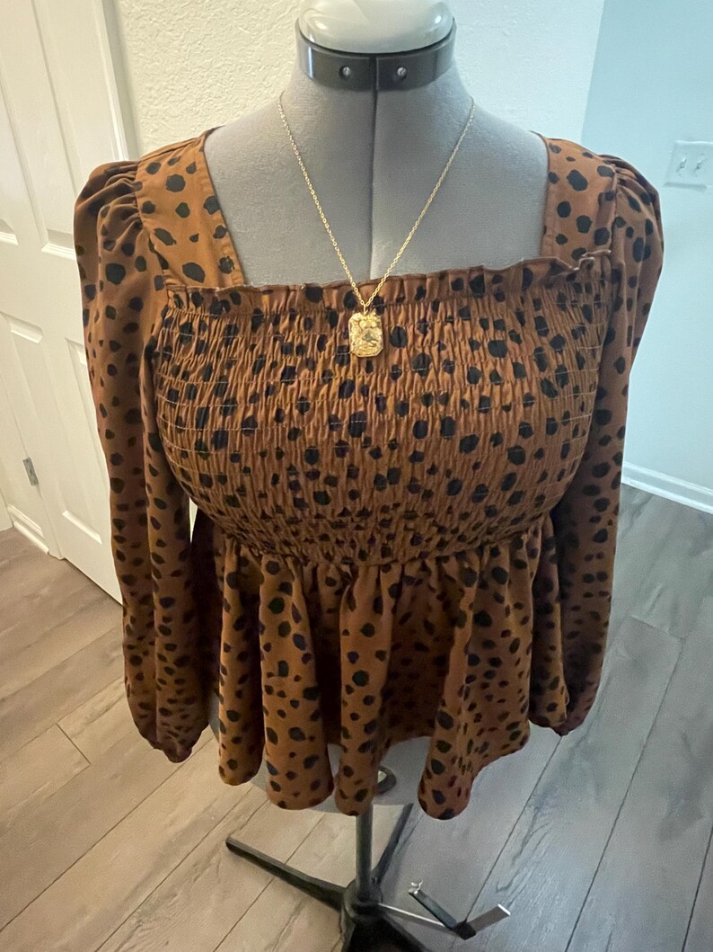 May include: A brown top with black polka dots, a square neckline, and long sleeves. The top has a smocked bodice and a peplum hem. A gold-coloured necklace with a square pendant hangs on the mannequin.
