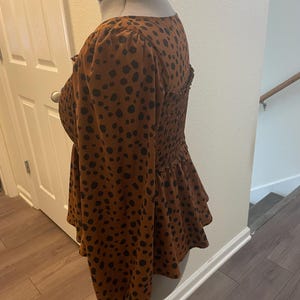 May include: A brown top with black polka dots, featuring long sleeves and a peplum hem. The top has a square neckline and a fitted, smocked bodice. The fabric is a soft, lightweight material.