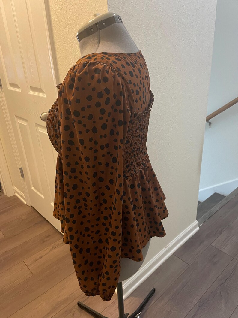 May include: A brown top with black polka dots, featuring long sleeves and a peplum hem. The top has a square neckline and a fitted, smocked bodice. The fabric is a soft, lightweight material.