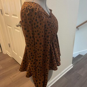 May include: A brown blouse with black polka dots. The long-sleeved top features a peplum hem and a smocked bodice. The blouse is displayed on a mannequin.