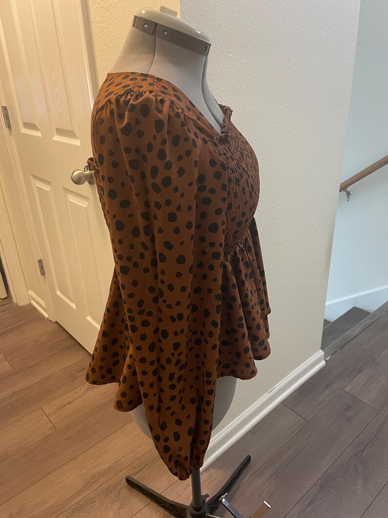 May include: A brown blouse with black polka dots. The long-sleeved top features a peplum hem and a smocked bodice. The blouse is displayed on a mannequin.