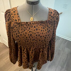 May include: A brown top with black polka dots, long sleeves, and a square neckline. The top has a smocked bodice and a ruffled peplum. A gold pendant necklace hangs on the mannequin.