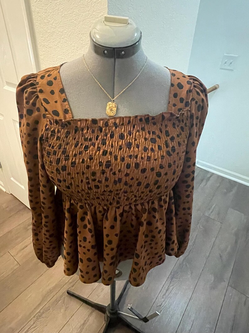 May include: A brown top with black polka dots, long sleeves, and a square neckline. The top has a smocked bodice and a ruffled peplum. A gold pendant necklace hangs on the mannequin.