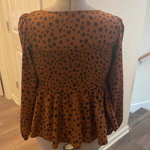 May include: A brown blouse with black polka dots, long sleeves, and a square neckline. The bodice is gathered, and the hem is a peplum style. The garment is displayed on a mannequin.