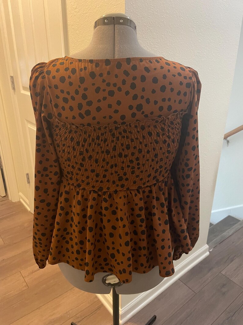 May include: A brown blouse with black polka dots, long sleeves, and a square neckline. The bodice is gathered, and the hem is a peplum style. The garment is displayed on a mannequin.
