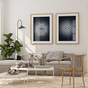 May include: Two framed wall art prints with geometric designs hang above a gray sofa. The prints have gold frames and feature intricate patterns in shades of blue and white against a dark background. The room is decorated with a large potted plant and a modern lamp.