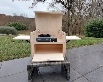 May include: A light-colored wooden camping kitchen with a gas stove, side tables, and storage. The unit is open, revealing the stove and interior space. It is set up outdoors on a patio, ready for cooking.