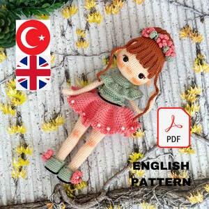 May include: Crochet doll pattern in PDF format. The doll is wearing a pink skirt, green top, and has brown hair with pink flowers. The pattern is available in English.