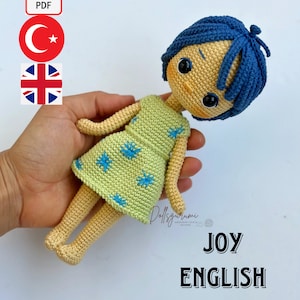 以下が含まれることがあります： 英語のクロシェドールパターン。青い髪の女の子が緑の星柄のドレスを着ています。パターンはPDFダウンロードで入手できます。