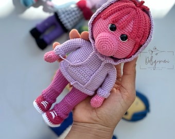 Könnte beinhalten: Ein gehäkeltes Plüschtier in Form eines Schweins, das einen lila Hoodie und eine rosa Hose trägt. Das Spielzeug hat eine rosa Nase und große schwarze Augen. Das Spielzeug wird in einer Hand gehalten.
