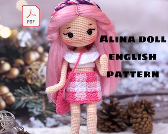 Puede incluir: Un patrón de muñeca de crochet para una muñeca rosa con una falda a cuadros blancos y rosas, un top blanco y cabello rosa. El patrón se llama "Alina Doll English Pattern" y está disponible para descargar en PDF.