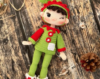 Puede incluir: Una muñeca de elfo de crochet con ropa verde y roja, cabello castaño y una cara guiñando un ojo. La muñeca está tumbada sobre una superficie de madera.