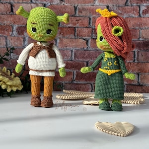 Puede incluir: Dos muñecos de ganchillo hechos a mano, uno parecido a Shrek y otro a Fiona, están uno al lado del otro. Shrek es verde con una camisa blanca, un chaleco marrón y pantalones naranjas. Fiona lleva un vestido verde y pelo rojo. Los muñecos están sobre una superficie blanca con un fondo de ladrillos.