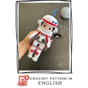 May include: A crocheted astronaut doll in a white spacesuit with red and blue accents. The doll has a white helmet with a red stripe and a blue antenna. The text "CROCHET PATTERN IN ENGLISH" is displayed below the doll.