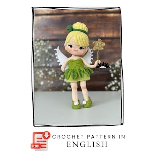 Puede incluir: Una muñeca de hada de ganchillo con un vestido, zapatos y moño verdes, sosteniendo una varita de estrella dorada. La muñeca tiene alas blancas y una expresión dulce. Incluye el texto "CROCHET PATTERN IN ENGLISH".