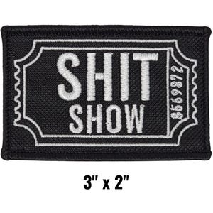 May include: A black embroidered patch with the words "SHIT SHOW" in white, framed by a white decorative border. The patch measures 3 inches by 2 inches.