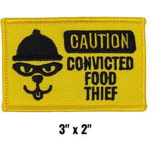May include: A yellow embroidered patch with black text and a cartoon image of a thief wearing a hat and eye mask. The text reads "CAUTION CONVICTED FOOD THIEF". The patch measures 3" x 2".