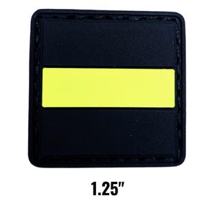 May include: A black square patch with a bright yellow horizontal stripe. The patch measures 1.25 inches across. The edges are stitched with black thread. The patch is designed for application on clothing or gear.