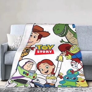 May include: A white fleece blanket featuring a colorful Toy Story graphic print. The design includes characters Woody, Buzz Lightyear, Jessie, Rex, an Alien, Forky, Ducky, and Bunny, with the red and yellow Toy Story logo centered on the fabric.