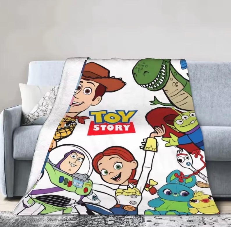 May include: A white fleece blanket featuring a colorful Toy Story graphic print. The design includes characters Woody, Buzz Lightyear, Jessie, Rex, an Alien, Forky, Ducky, and Bunny, with the red and yellow Toy Story logo centered on the fabric.