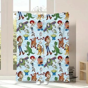 May include: A light blue fleece blanket featuring a repeating pattern of Toy Story characters including Woody, Buzz Lightyear, Jessie, Rex, Slinky Dog, Mr. Potato Head, Forky, and the green aliens against a backdrop of white clouds.