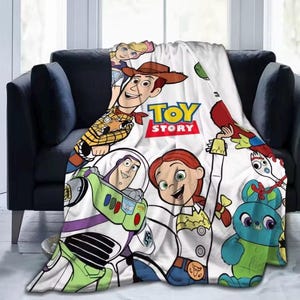 May include: A white fleece blanket featuring a colorful Toy Story graphic print. The design includes characters Woody, Buzz Lightyear, Jessie, Bo Peep, Forky, and Ducky. The central text reads TOY STORY in bold yellow and red lettering.