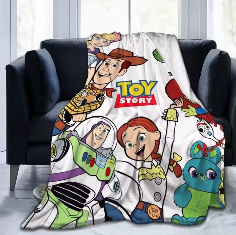 May include: A white fleece blanket featuring a colorful Toy Story graphic print. The design includes characters Woody, Buzz Lightyear, Jessie, Bo Peep, Forky, and Ducky. The central text reads TOY STORY in bold yellow and red lettering.