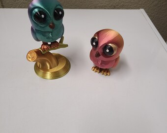 May include: Two small owl figurines, one perched on a gold branch and the other standing on a white surface. The owl on the branch is iridescent blue and green, while the other owl is a deep red with gold accents.