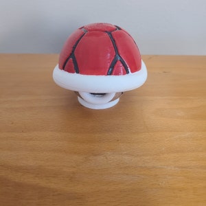 Puede incluir: Una cáscara roja y blanca impresa en 3D de la serie de videojuegos Super Mario Bros. La cáscara es un objeto común en el juego y se usa para atacar a los enemigos.