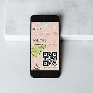 May include: A smartphone screen displaying a pink and white background with a margarita glass and a QR code. The text on the screen reads "Buy a Margarita for the Mrs." and "@venmo.user".