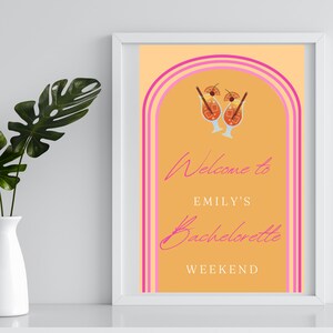 Peut inclure: Une pancarte de bienvenue pour un enterrement de vie de jeune fille avec un fond p&ecirc;che et des rayures roses. La pancarte indique "Bienvenue &agrave; Emily's Bachelorette Weekend" avec deux cocktails au centre.