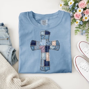 Może przedstawiać: Jasnoniebieska koszulka Comfort Colors z patchworkowym wzorem krzyża. Krzyż wykonany jest z różnych tkanin dżinsowych w odcieniach niebieskiego, różowego i białego. Koszulka jest złożona na białej powierzchni, z dżinsami, swetrem i białymi trampkami w tle.